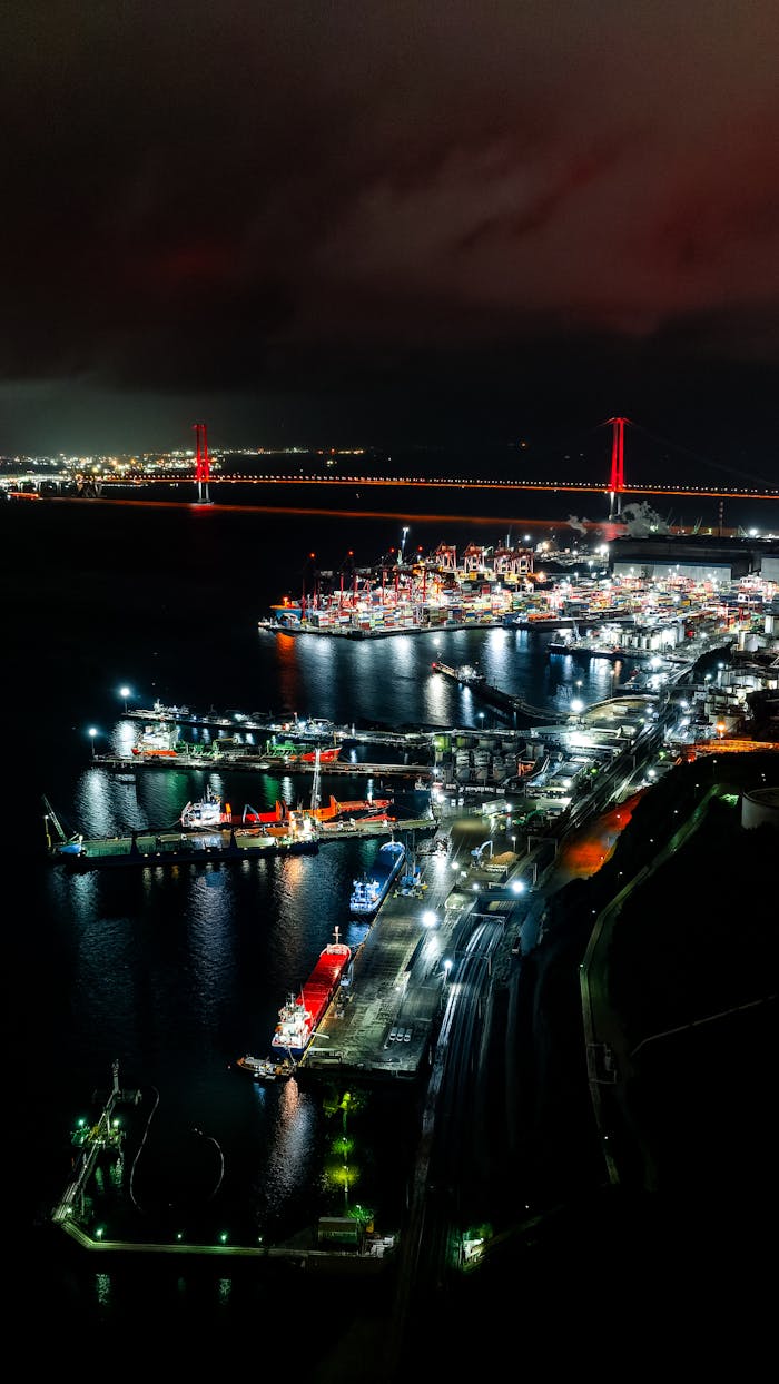 Aerial night view of a vibrant urban port with illuminated ships and city lights reflecting on water.